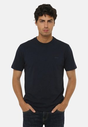 Uomo con capelli scuri e ricci che indossa una maglietta nera semplice a maniche corte e jeans scuri con le mani nelle tasche, espressione neutra.