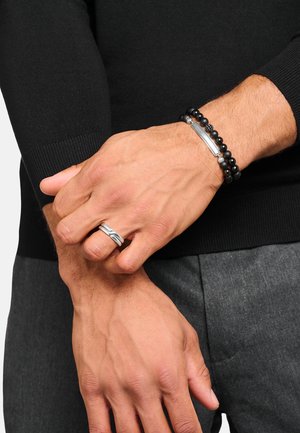 Hände mit einem silbernen gedrehten Ring am linken Ringfinger und zwei schwarzen Perlenarmbändern am rechten Handgelenk über einem schwarzen Langarmshirt.