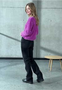 Stickad, livfull rosa tröja med texturerad yta, kombinerad med svarta jeans och svarta ankelboots, mot en betongvägg och enkel inredning.