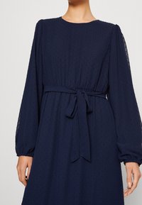 Granatowa sukienka z okrągłym dekoltem, bufkami na długich rękawach, teksturowanymi kropkami oraz materiałowym paskiem wiązanym w talii dla dopasowanej sylwetki.