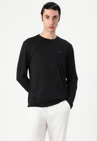 Zwart T-shirt met lange mouwen van zachte stof met een ronde hals en een klein logo op de linkerkant van de borst, gecombineerd met witte broek.