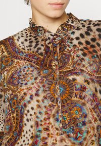 Gros plan sur une personne portant un blouse à motifs avec des couleurs vives et des designs complexes, présentant un col volanté et un nœud au cou.