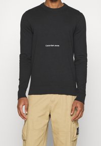 Svart bomullströja med långa ärmar, ribbade muddar och fåll, med en vit "Calvin Klein Jeans"-logotyp mitt fram.