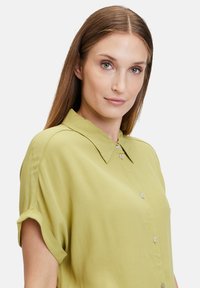 Short-sleeve, collared shirt in light green fabric. Features buttons down the front and rolled cuffs. Soft, smooth texture.