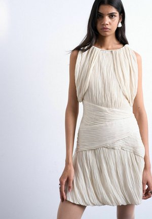 Femme aux longs cheveux foncés portant une robe sans manches plissée de couleur crème avec un détail superposé à la taille, debout devant un fond clair uni.