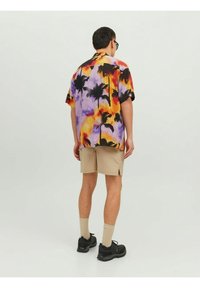 Camisa de manga corta con un estampado colorido de palmeras en un degradado de naranja, púrpura y amarillo. Combinada con pantalones cortos beige y zapatos negros.