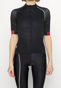 Femme portant un maillot de cyclisme noir à manches courtes avec des poignets rouges et un short de cyclisme noir rembourré assorti, avec une bande latérale blanche.