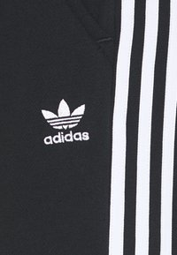 Černé sportovní kalhoty se třemi vertikálními bílými pruhy a bílým logem Adidas. Materiál je hladký s mírným leskem.