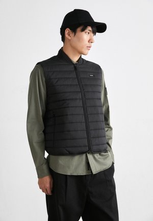 Gilet imbottito nero con chiusura a zip, design trapuntato e etichetta sul petto sinistro; indossato sopra una camicia verde e abbinato a un berretto nero.