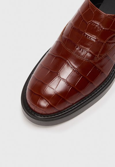 Zapato de cuero marrón con grabado de cocodrilo, con puntera redondeada, superficie texturizada y suela de goma negra con detalles acanalados.