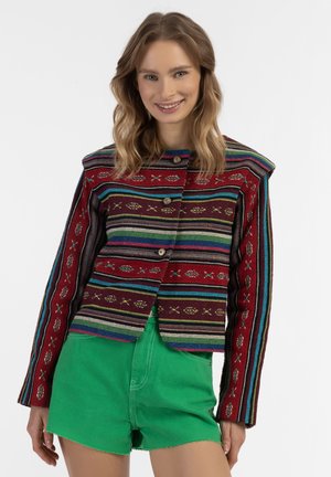 Rode gestreepte jas met een patroon, gekenmerkt door veelkleurige strepen, geborduurde accenten en houten knopen. Gepaarde met groene shorts.