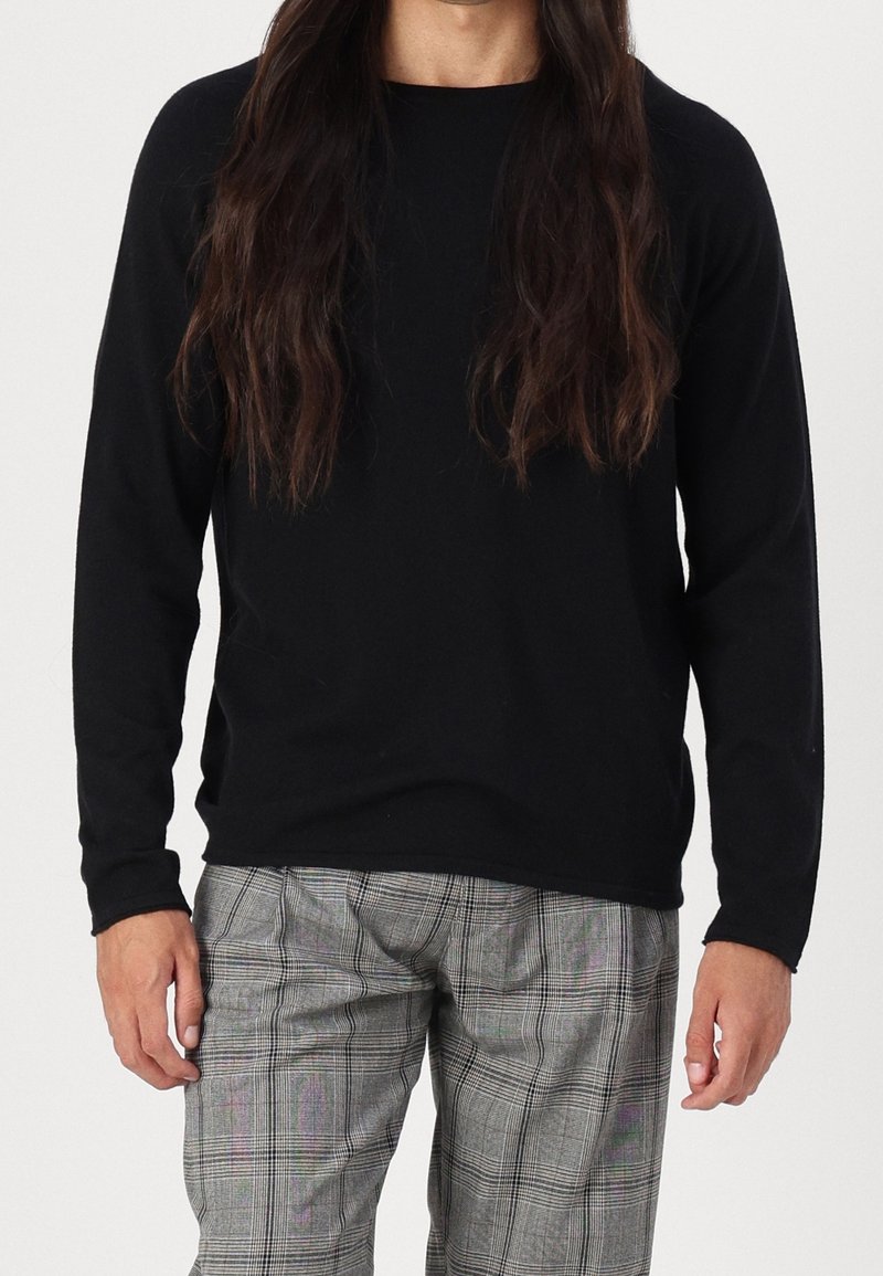Homme portant une chemise noire à manches longues et un pantalon à carreaux gris, debout avec les bras détendus le long du corps devant un fond uni.