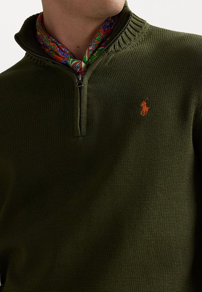 Olivengrøn strikket pullover med halv-zip krave, prydet med ribbede detaljer og et orange broderet logo på brystet. Farverig mønstret tørklæde synlig.