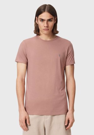 Junger Mann mit schulterlangen Haaren, der ein einfaches, staubiges rosa kurzärmeliges T-Shirt und hellbeige Hose trägt, vor einem schlichten Hintergrund stehend.
