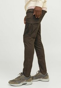 Cargo-broek in donkerbruin katoenmix, met zijzakken, taps toelopende benen en stikdetails. Gecombineerd met grijze sneakers.