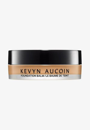 Kevyn Aucoin Foundation Balm in a round, black lid container. The base is tan with a smooth, creamy texture. Text reads "Foundation Balm."