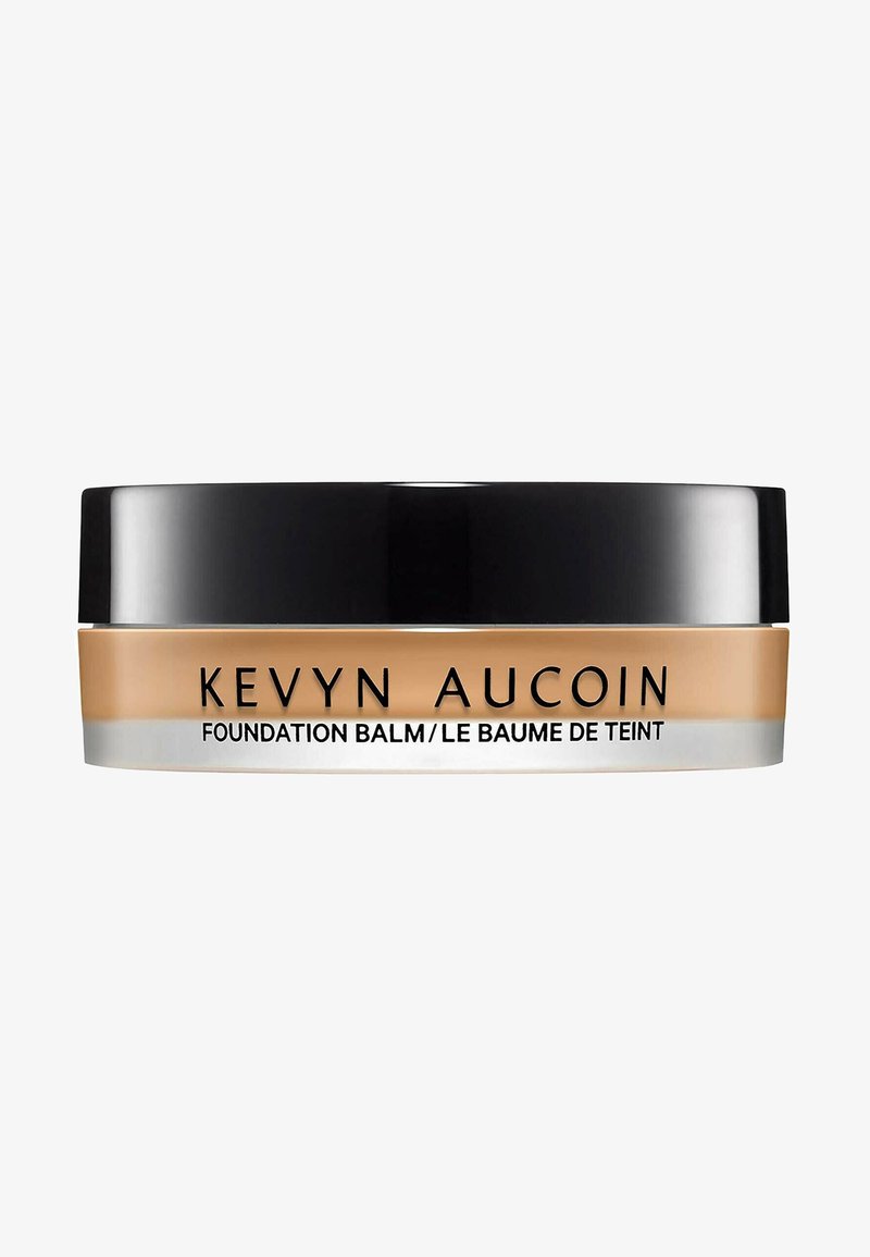 Kevyn Aucoin Foundation Balm in a round, black lid container. The base is tan with a smooth, creamy texture. Text reads "Foundation Balm."