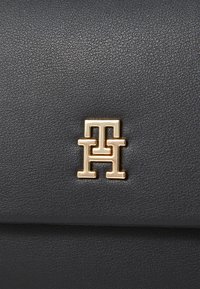 Bolso de mano de cuero negro con una prominente placa de logo en tono dorado con letras entrelazadas en la parte frontal. Textura suave con un diseño minimalista.