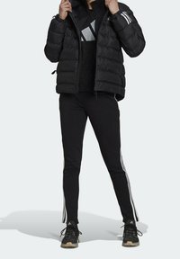 Doudoune noire matelassée horizontale, associée à un pantalon de survêtement noir avec des bandes blanches sur les côtés, et des chaussures de sport noires.