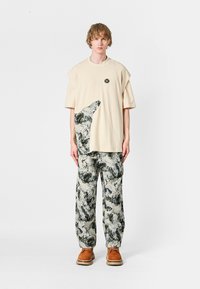 Joven vestido con una camiseta oversized beige con un parche abstracto en blanco y negro, pantalones a juego con un patrón abstracto y zapatos marrones.