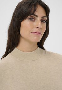 Beige højhalsstriktrøje lavet af blødt strikket materiale, med ribbet krave og en glat, jævn tekstur.