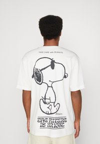 T-shirt en coton blanc avec un grand graphique noir de Snoopy au dos, accompagné du texte "L'AVENIR EST PROMETTEUR" en dessous. Manches courtes.