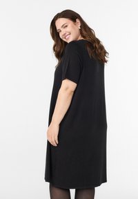 Hymyilevä nainen, jolla on pitkät ruskeat hiukset, hopeiset korvakorut ja löysä musta lyhythihainen mekko, seisomassa valkoista, yksinkertaista taustaa vasten.