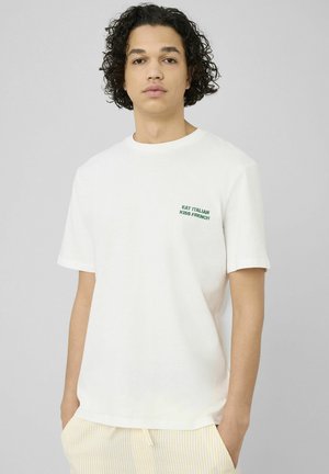 Jeune personne aux cheveux bouclés portant un t-shirt blanc avec un texte vert « EAT ITALIAN KISS FRENCH » et un pantalon clair à rayures, les mains dans les poches.