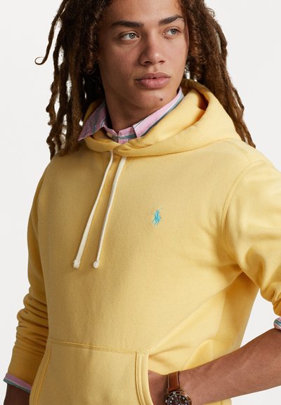 Rumena bombažna kapuca z žepom spredaj, modrologo na prsih in vrvico, nosena čez svetlo roza in modro srajco s ovratnikom.