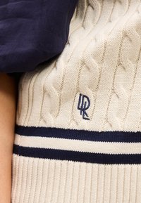 Κοντινό πλάνο κρεμ πουλόβερ με πλεκτό σχέδιο καλώδιο, με μπλε ναυτικό ρίγες και κεντημένα αρχικά «LR» κοντά στο στήθος. Μπλε ναυτικό ύφασμα μερικώς ορατό στον ώμο.