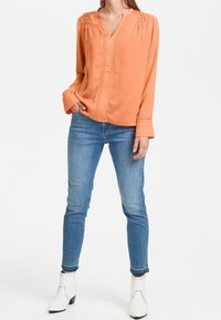 Blusa arancione a maniche lunghe con scollatura a V, dettagli arricciati sulle spalle e una cucitura frontale singola, abbinata a jeans blu aderenti e stivaletti bianchi alla caviglia.