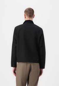 Chaqueta negra sin cuello con cierre abotonado y lados ajustables. Textura suave, corte entallado y ligero brillo. Diseño sencillo, sin patrones.