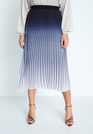 Donna che indossa una gonna midi plissettata con una sfumatura ombré dal blu navy scuro al bianco, abbinata a tacchi metallici con cinturini e un maglione leggero.