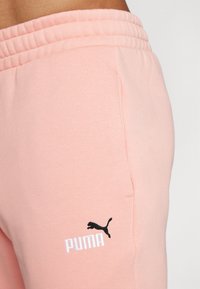 Gaiši rozā kokvilnas treniņu bikses ar ribotu jostu, sānu kabatu un melnu izšuvumu Puma logotipu ar baltu tekstu.