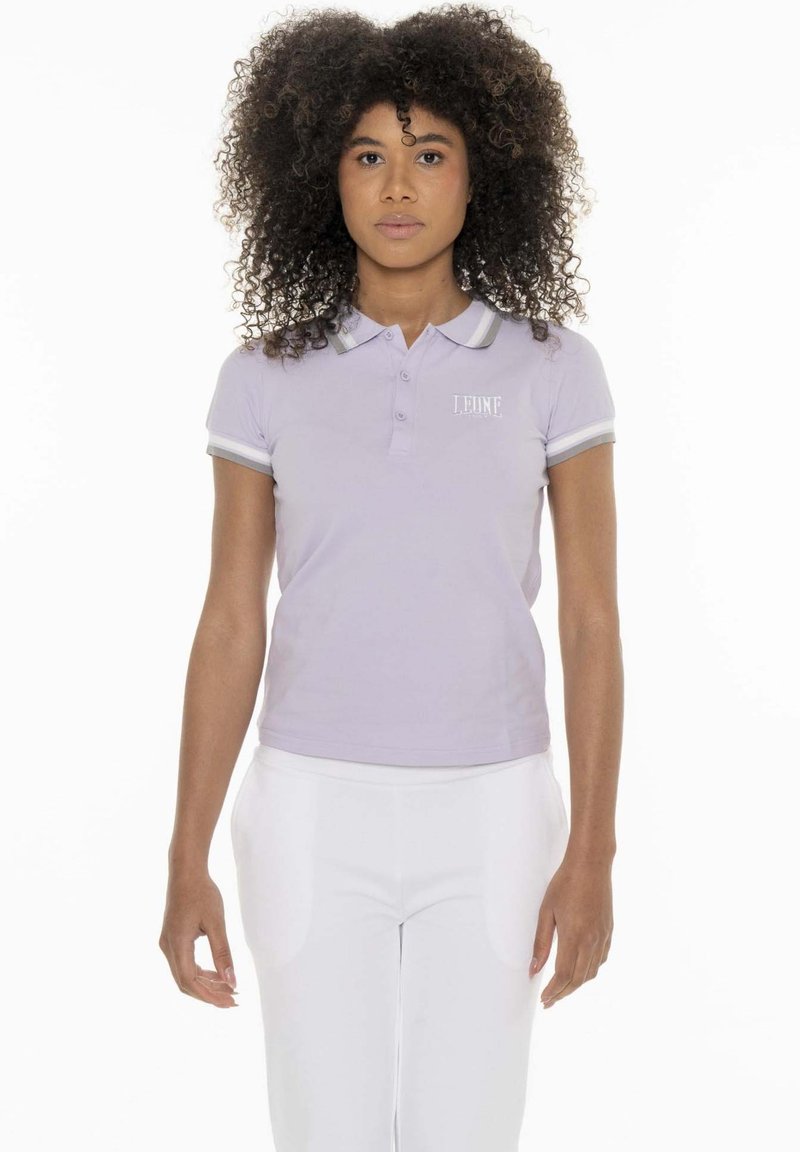 Femme aux cheveux bouclés, debout face à l'avant, portant un polo violet clair et un pantalon blanc sur un fond blanc.