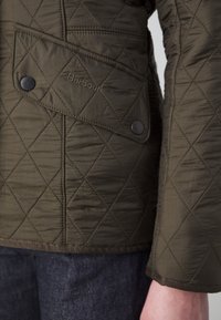 Poche cousue matelassée vert olive de la veste Barbour avec des boutons-pression et des coutures visibles sur la manche et le corps, portée sur un pantalon foncé.