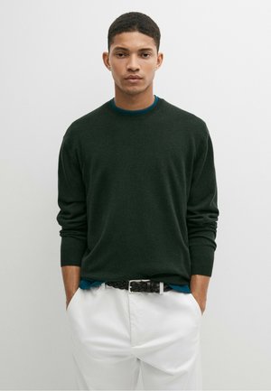 Uomo che indossa un maglione verde scuro, camicia blu sotto, pantaloni bianchi e cintura nera intrecciata con le mani in tasca su sfondo uniforme.