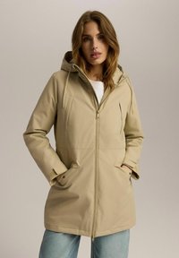Beżowa wodoodporna kurtka z kapturem, z przednim zamkiem błyskawicznym i dwiema bocznymi kieszeniami. Charakteryzuje się gładką, teksturowaną tkaniną i regulowanymi sznurkami.