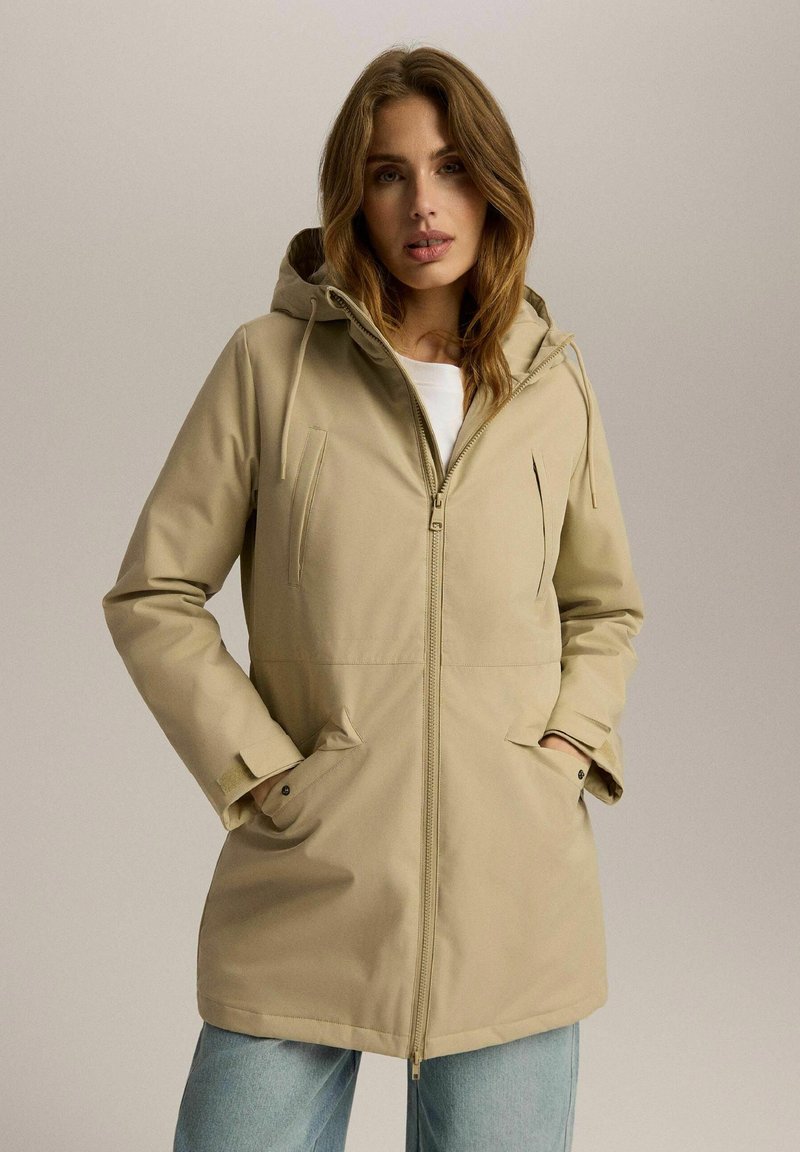 Beżowa wodoodporna kurtka z kapturem, z przednim zamkiem błyskawicznym i dwiema bocznymi kieszeniami. Charakteryzuje się gładką, teksturowaną tkaniną i regulowanymi sznurkami.