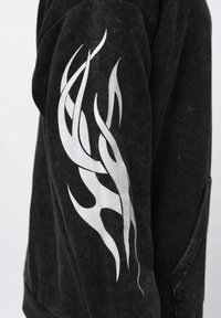 Sweatshirt preto em tecido texturizado; apresenta um destacável desenho de chama branca na manga esquerda e um bolso lateral.