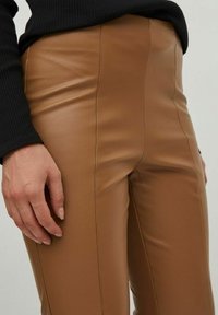 Pantalons en faux cuir marron taille haute avec des coutures décoratives et une texture lisse, associés à un haut noir à manches longues côtelé.