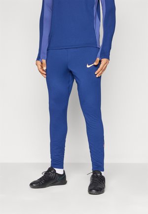 Persoon die blauwe Nike sportbroek en bijpassende long-sleeve top draagt, met zwarte sportschoenen, staand tegen een effen achtergrond.