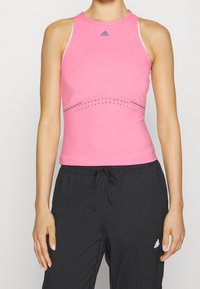 Różowy bezrękawnik z wysokim kołnierzem, z detalami wentylacyjnymi i szarą linią akcentową na klatce piersiowej. Zestawiony z czarnymi spodniami z elastyczną talią.