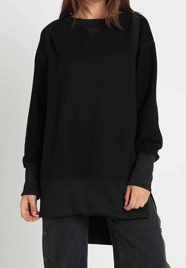 Jeune personne portant un sweat-shirt noir surdimensionné à manches longues et un pantalon large sombre avec des boutons sur le côté.
