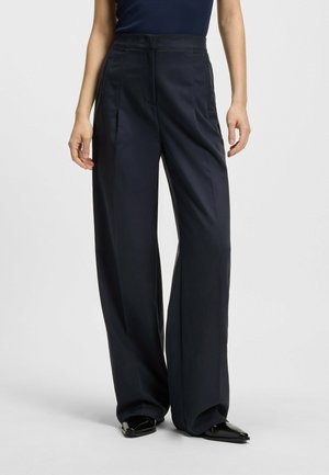 Marineblaue weit geschnittene Hose mit glatter Textur, ausgestattet mit Vorderfalten und hoher Taille. Kombiniert mit schwarzen spitzen Schuhen.