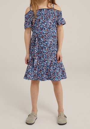 Pige iført en blå, blomstret kjole med bare skuldre og bånd i taljen samt beige slip-on sko, stående foran en ensfarvet baggrund.