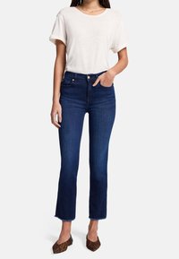 Mujer vistiendo una blusa blanca de manga corta, unos jeans de tiro alto de color azul oscuro con dobladillos deshilachados y unos zapatos planos de punta con estampado de leopardo, con una mano en el bolsillo.