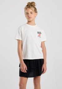 Weißes T-Shirt mit kurzen Ärmeln, das ein Wassermelonenmotiv und schwarzen Text auf der linken Brust zeigt, kombiniert mit einem schwarzen mehrlagigen Volantrock.