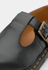 Chaussure en cuir noir avec bout rond, design à bride en T, fermeture à boucle, détail découpé, intérieur marron et surpiqûres jaunes contrastantes le long de la semelle.