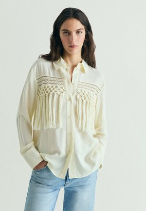 Mujer de cabello largo y oscuro, vestida con una camisa crema abotonada con flecos y detalles de encaje, combinada con jeans azul claro, de pie con las manos en los bolsillos.